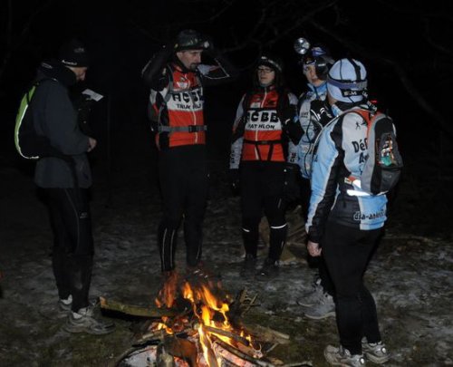 Coureurs en tenue de raid et lampe frontale rassemblés de nuit autour d’un feu de camp sur un sol enneigé